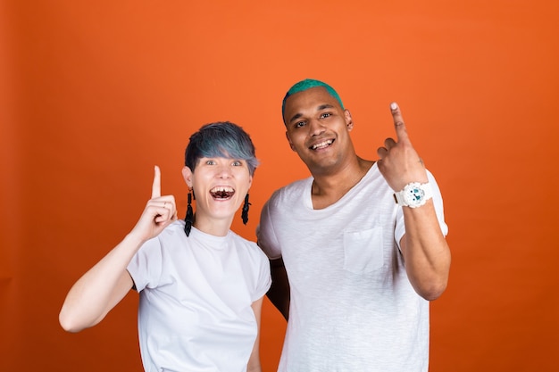 Hombre y mujer joven en blanco casual sobre pared naranja, ambos sonrientes felices apuntan hacia arriba con el dedo índice