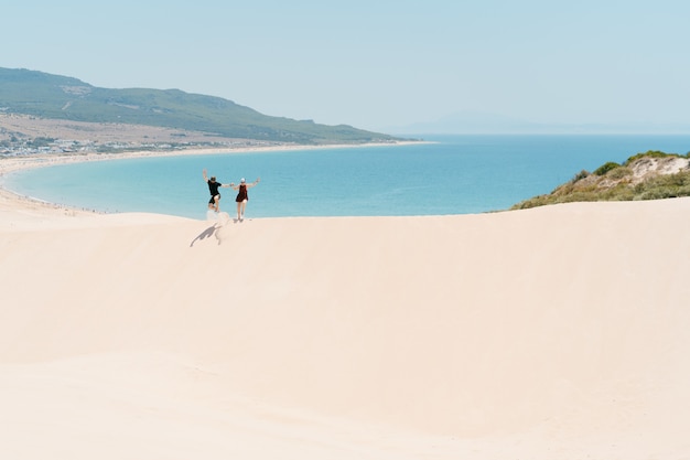 Hombre y mujer joven saltando en las dunas de arena con vistas al concepto de playa de vacaciones y libertad