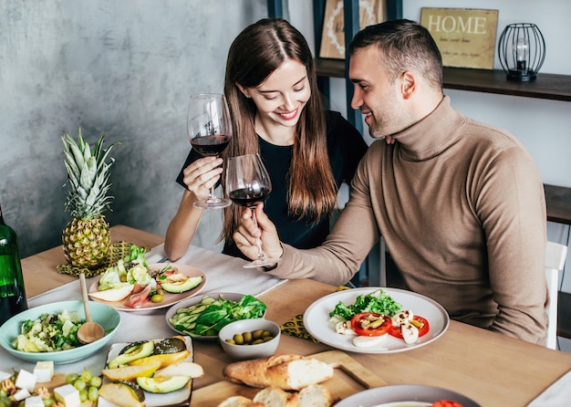 El hombre y la mujer jóvenes están sentados en una mesa cubierta con comida y bebidas con vasos de vino en sus manos