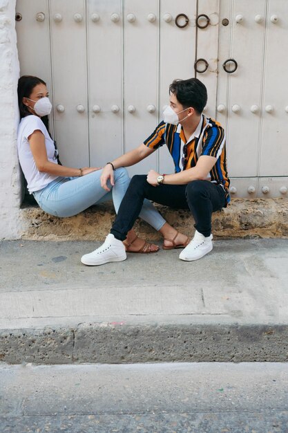 Un hombre y una mujer jóvenes con una máscara desechable vestidos casualmente hablando mientras están sentados al aire libre