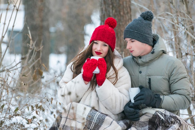 El hombre y la mujer jóvenes se sientan abrazándose unos a otros envueltos en una manta y bebiendo té de tazas en un parque de invierno cubierto de nieve. Cita romántica en el bosque de invierno.