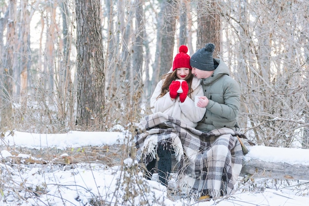 El hombre y la mujer jóvenes se sientan abrazándose en un parque de invierno cubierto de nieve, envueltos en una manta y bebiendo té de tazas. Cita romántica en el bosque de invierno.