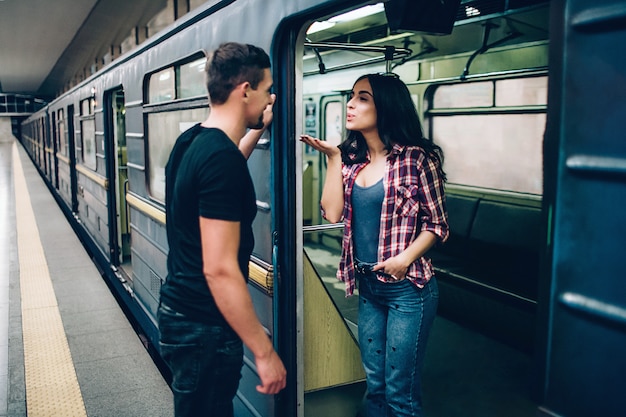Hombre y mujer jóvenes usan bajo tierra. Pareja en metro Encantadora joven enviar besos al hombre. Guy la mira y sonríe. Juntos solos en plataforma y carro subterráneo.