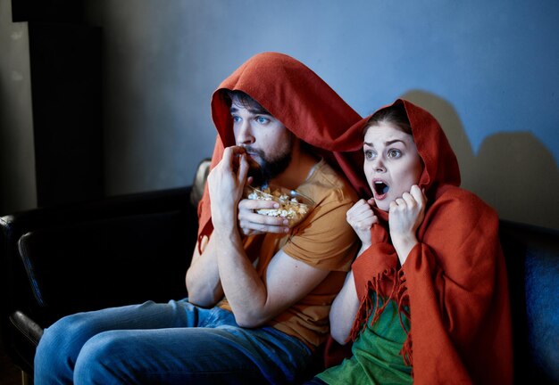 Un hombre y una mujer con una manta roja en la cabeza están sentados en el sofá frente al televisor en el interior