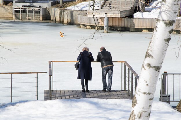 Un hombre y una mujer de mediana edad se paran en el puente junto al estanque cubierto de hielo y admiran a los patos.