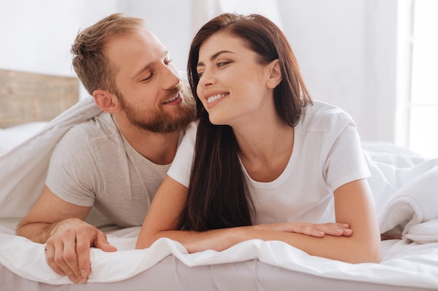 Hombre y mujer milenarios sonriendo ampliamente mientras están acostados en la cama y hablando sobre sus relaciones