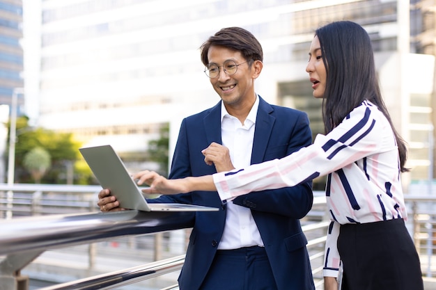 Hombre y mujer de negocios asiáticos trabajando en equipo portátil de pie fuera del edificio de oficinas.