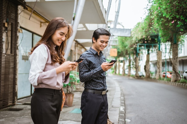 Hombre y mujer, posición, en, un, acera, sonriente, utilizar, el suyo, teléfono