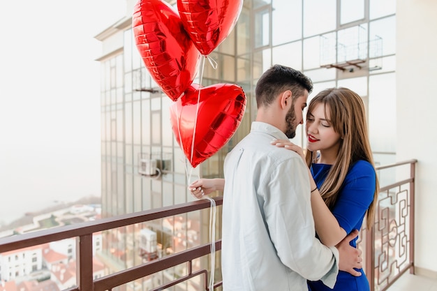 Hombre y mujer con regalo y globos rojos en forma de corazón en el balcón en casa