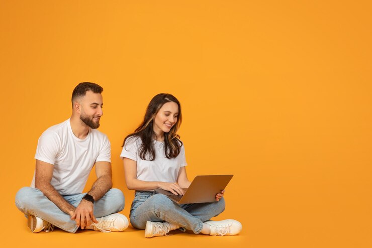 Un hombre y una mujer relajados sentados con las piernas cruzadas en el suelo con la mujer usando una computadora portátil