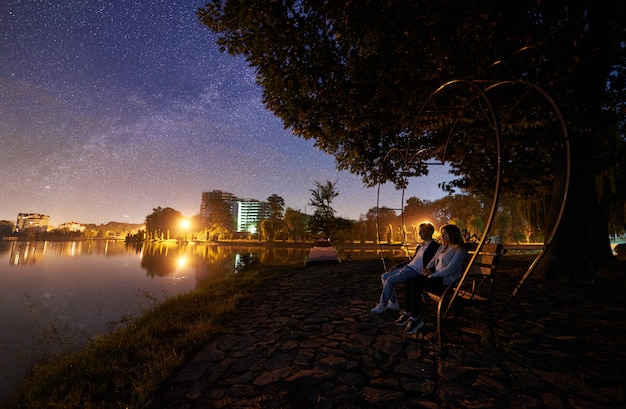 Hombre y mujer sentada en el banco en la orilla cerca del lago bajo el árbol. Pareja disfrutando de la vista del cielo nocturno lleno de estrellas, vía láctea, superficie del agua tranquila, luces de la ciudad en el fondo. Concepto de estilo de vida al aire libre