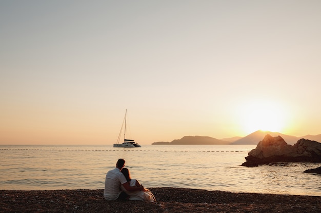 Un hombre y una mujer se sientan abrazados en la orilla de un mar en calma y miran el velero