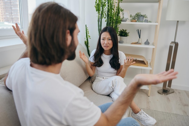 Un hombre y una mujer se sientan en el sofá de casa con camisetas blancas y gritando se comunican escandalosamente no se entienden Una pelea en la familia de dos cónyuges y agresión emociones tristes