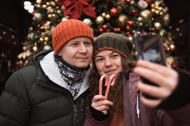 Hombre y mujer con sombreros y ropa de invierno tomando selfie por teléfono en el fondo del árbol de navidad