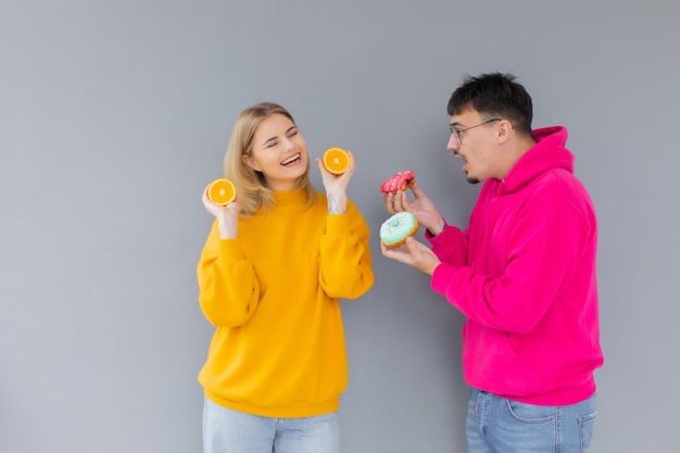 Hombre y mujer sosteniendo donuts y naranja sobre un fondo gris Concepto de estilo de vida saludable