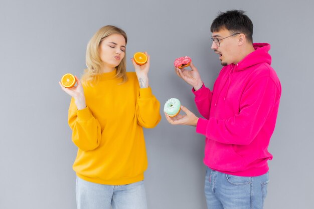 Hombre y mujer sosteniendo donuts y naranja sobre un fondo gris Concepto de estilo de vida saludable