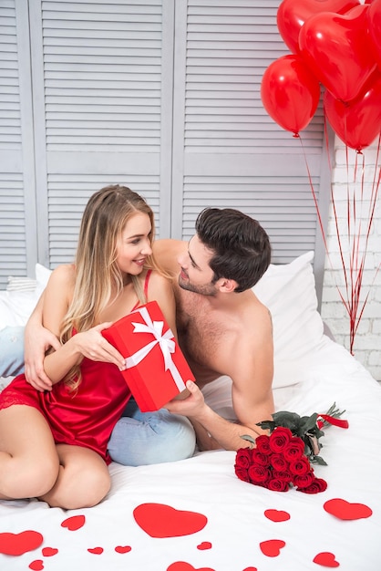 Hombre y mujer sosteniendo una gran caja de regalo roja y mirándose mientras están sentados en la cama concepto de día de san valentín