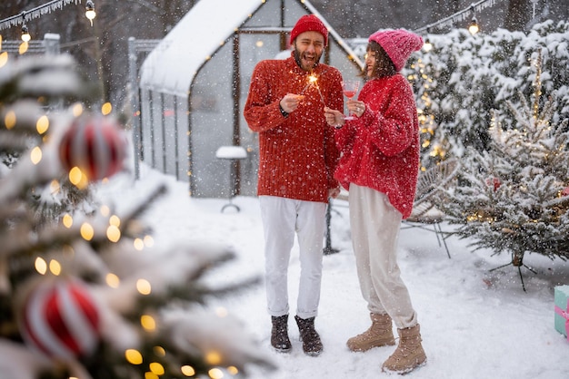 Hombre y mujer con suéteres rojos celebran las vacaciones de Año Nuevo encendiendo chispas y divirtiéndose cerca del árbol de Navidad en el patio trasero nevado