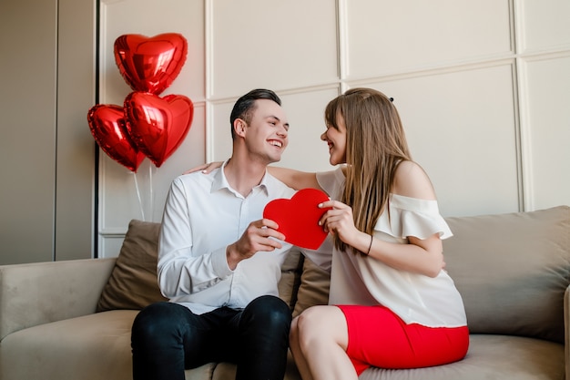 Hombre y mujer con tarjeta de san valentín en forma de corazón rojo y globos en casa en el sofá