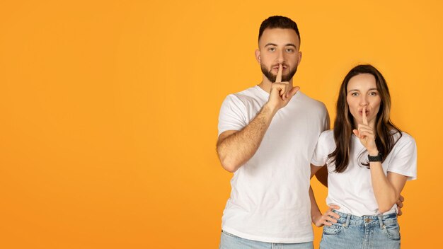 Hombre y mujer tranquilos y confiados en camisas blancas colocando los dedos en los labios en un gesto de silencio