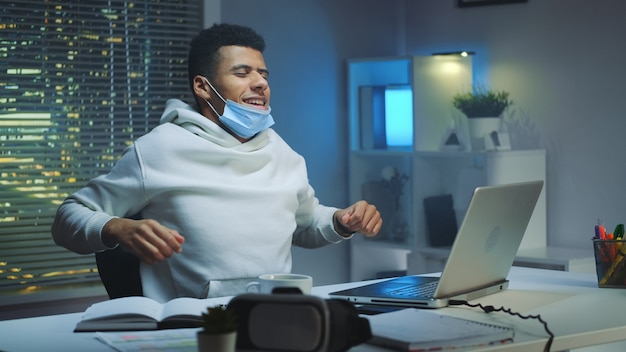 Hombre multiétnico cansado con máscara protectora trabajando en casa tarde en la noche y haciendo actividades de calentamiento