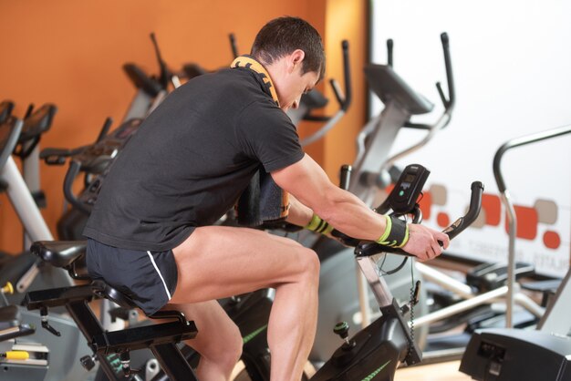 Hombre musculoso andando en bicicleta en el gimnasio, ejercitando las piernas haciendo ejercicios cardiovasculares, andando en bicicleta.