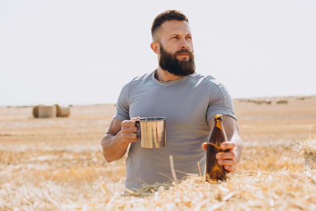 Un hombre musculoso barbudo vierte y prueba cerveza fresca en un campo de trigo en medio de balas