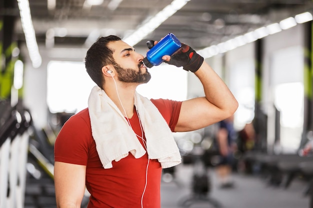 Hombre musculoso barbudo viste camiseta roja bebe agua de una botella azul en el gimnasio