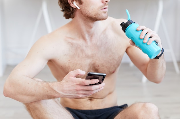 Hombre musculoso sin camisa bebiendo agua y usando su teléfono móvil después del entrenamiento deportivo