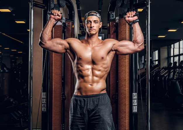 Un hombre musculoso sin camisa está haciendo ejercicios con aparatos de entrenamiento en un club de gimnasia oscuro.