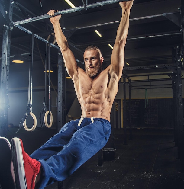 Hombre musculoso sin camisa en pantalones deportivos azules haciendo ejercicios en un gimnasio en barra horizontal.