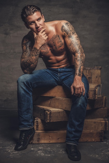 Hombre musculoso sin camisa con tatuajes en el cuerpo sentado en cajas de madera.