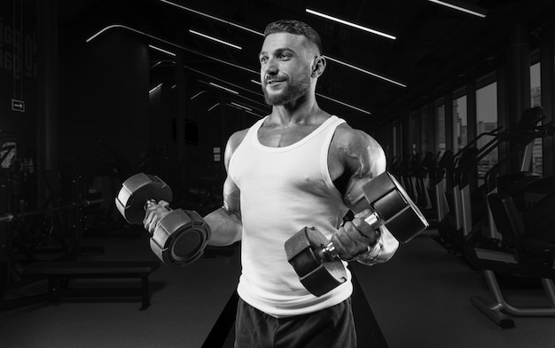 Hombre musculoso con una camiseta blanca trabaja en el gimnasio con pesas. Bombeo de bíceps. Concepto de fitness y culturismo.