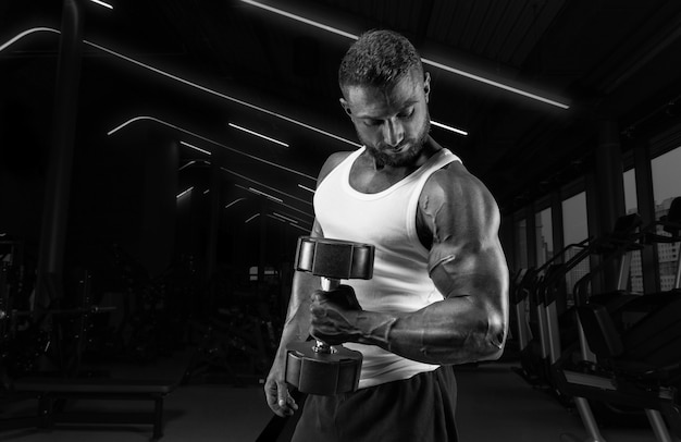 Hombre musculoso con una camiseta blanca trabaja en el gimnasio con pesas. Bombeo de bíceps. Concepto de fitness y culturismo.