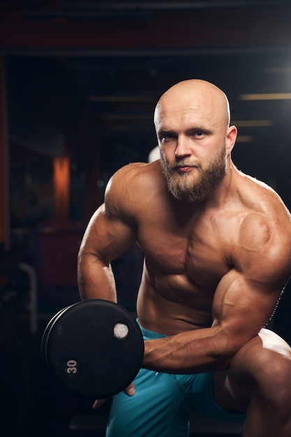 Hombre musculoso carismático trabajando sin camiseta y levantando pesas mientras mira a la cámara