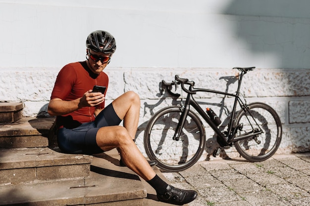 Hombre musculoso caucásico con ropa deportiva, casco y anteojos usando un teléfono inteligente moderno mientras se sienta cerca de una bicicleta al aire libre Concepto de recreación y tecnología de personas