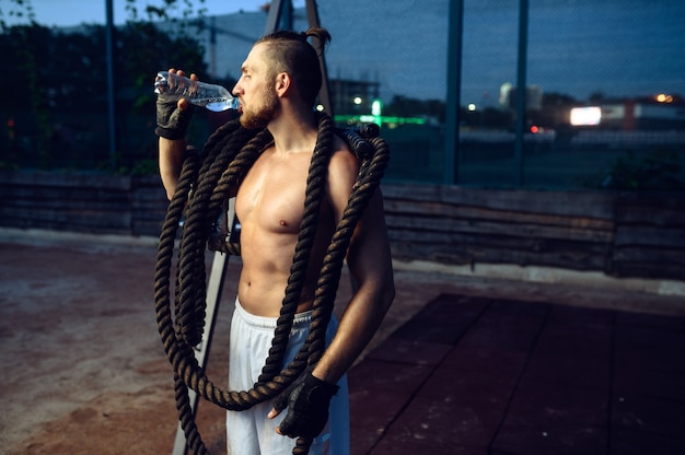 Hombre musculoso con cuerdas de crossfit bebe agua, entrenamiento en la calle