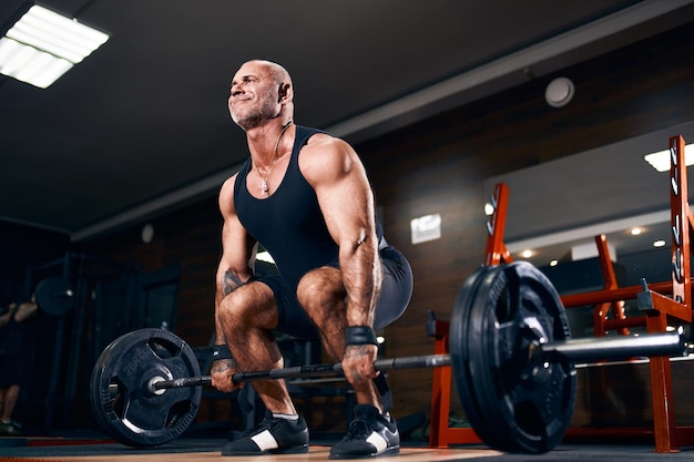 Hombre musculoso culturista haciendo ejercicio de peso muerto pesado con peso mientras está en el gimnasio sobre fondo oscuro