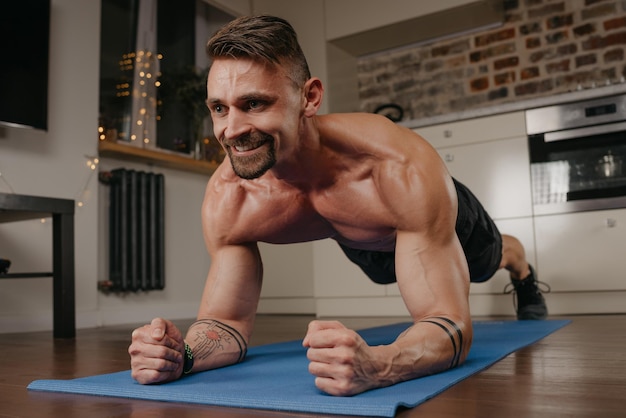 Un hombre musculoso feliz con el torso desnudo está haciendo una tabla en una estera de yoga azul en su apartamento por la noche Un tipo atlético sonriente con tatuajes en los antebrazos está entrenando en casa