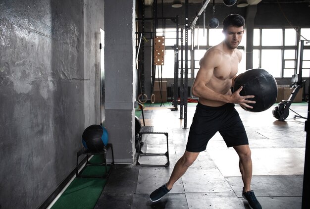 Hombre musculoso en forma concentrada sudoroso fuerte joven haciendo arrojar balón medicinal en la pared para entrenamiento crossfit entrenamiento duro en el gimnasio