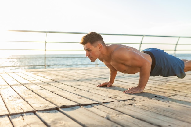 Hombre musculoso guapo fitness con torso desnudo haciendo flexiones en la playa al amanecer.