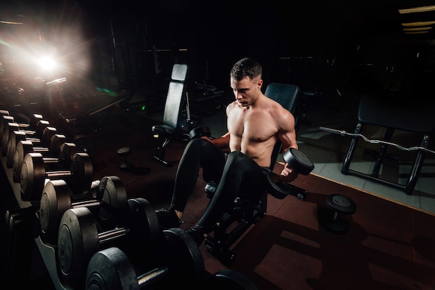 Hombre musculoso guapo haciendo ejercicio con pesas pesadas mientras está de pie en el gimnasio moderno oscuro