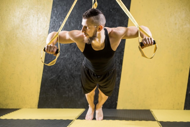 Foto hombre musculoso hace ejercicios con correas de fitness en el gimnasio con interior negro y amarillo