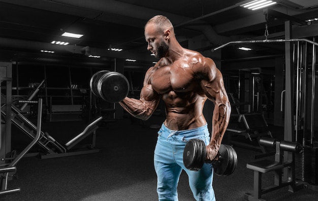 Hombre musculoso posa en el gimnasio en jeans con pesas en sus manos