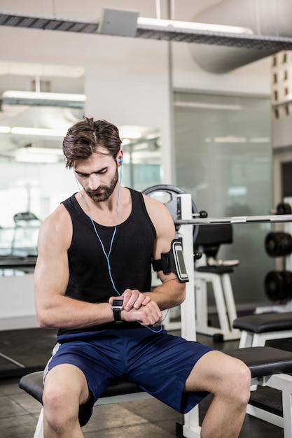 Hombre musculoso sentado en el banco con barra y usando smartwatch en el gimnasio