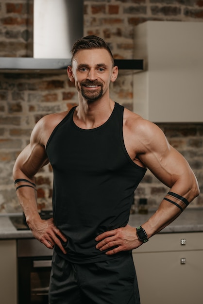 Un hombre musculoso sonriente con barba está posando con las manos en la cintura en su apartamento después de un entrenamiento