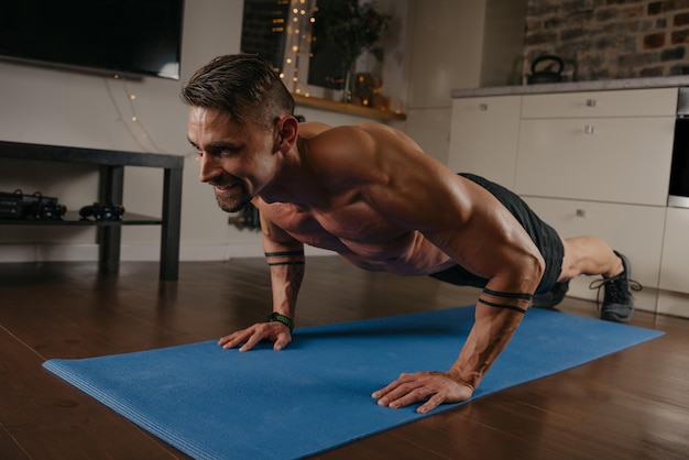 Un hombre musculoso con tatuajes está haciendo flexiones en una alfombra de yoga azul en su apartamento