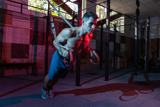 Hombre musculoso trabaja con el sistema trx en un gimnasio oscuro Atleta con entrenamiento de torso desnudo con correas de resistencia en luz de neón azul rojo Entrenamiento cruzado funcional