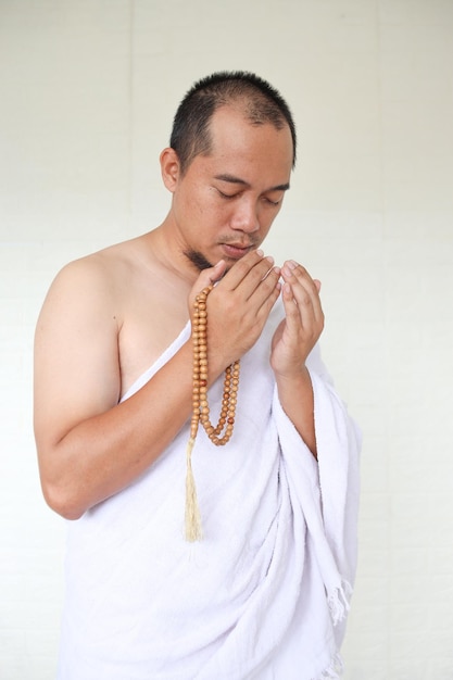 Hombre musulmán asiático vestido con ropa blanca de ihram y rezando mientras sostiene cuentas de oración