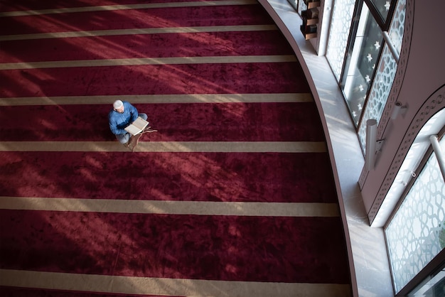 hombre musulmán rezando a Alá solo dentro de la mezquita y leyendo el libro islámico de acebo vista superior del Corán